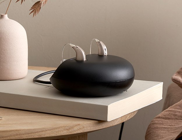 Two modern rechargeable hearing aids placed on a sleek black charging station resting on a book.