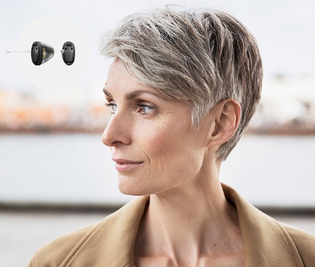 A short-haired woman is shown outdoors, wearing a tan coat. In the top-left corner, two black Oticon hearing aids are displayed on a white background, symbolizing advanced hearing solutions.