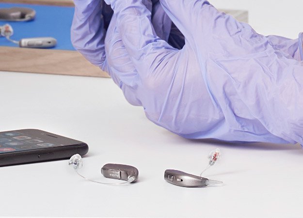 Hearing aids resting on a table next to a smartphone, with gloved hands handling equipment in the background.