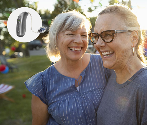 A sleek Oticon hearing aid is highlighted, positioned near two women embracing outdoors in a sunlit park-like setting, surrounded by trees, grass, and festive decorations.