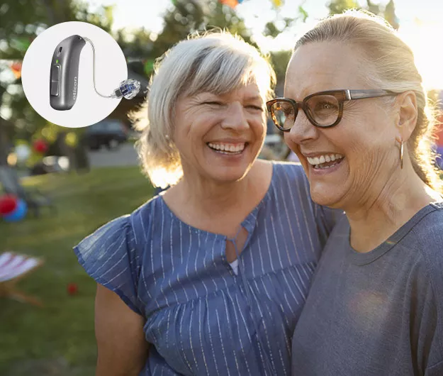 A sleek Oticon hearing aid is highlighted, positioned near two women embracing outdoors in a sunlit park-like setting, surrounded by trees, grass, and festive decorations.