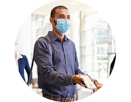 A man wearing a blue shirt and carrying a bag presents a document or phone to someone in a modern, well-lit building with large glass windows in the background.