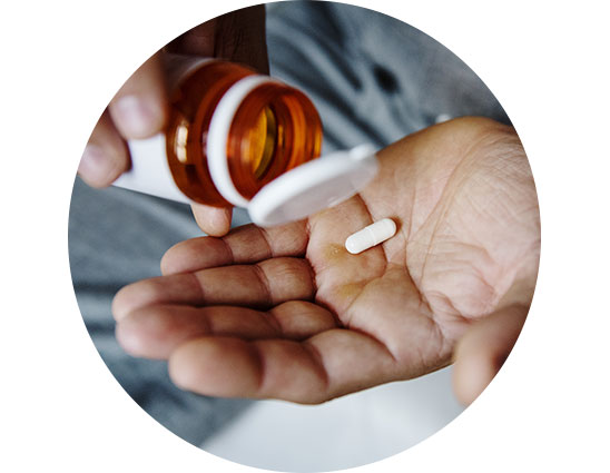 A hand holds a white capsule while an orange pill bottle is tipped, dispensing. The setting is neutral, suggesting a focus on health, medication, or treatment for wellness. No text is visible.