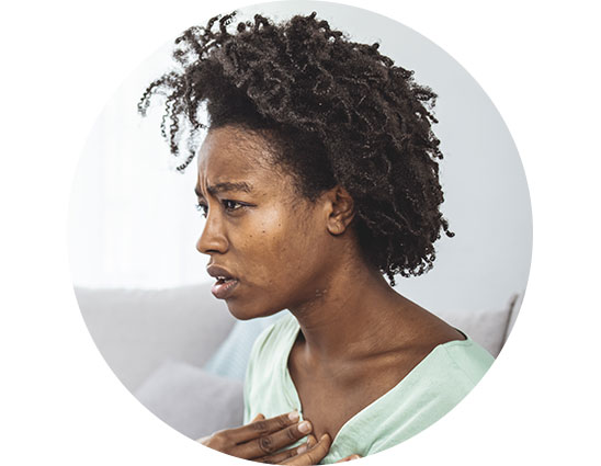 A person with natural curly hair is seated in a softly lit room wearing a light green top, placing one hand on their chest. The background includes a blurred sofa and cushions. No text or visible hearing aid is depicted.
