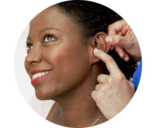 An audiologist adjusts a behind-the-ear hearing aid on a person, ensuring a proper fit. The close-up setting highlights professional hearing care and support in a clinical context. No text is visible.