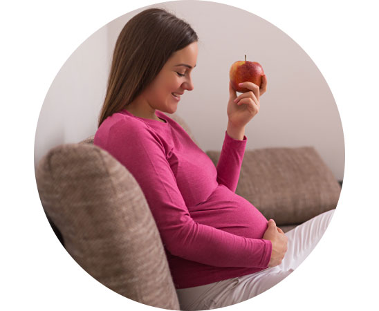 Pregnant woman in a pink shirt sitting on a sofa holding an apple.