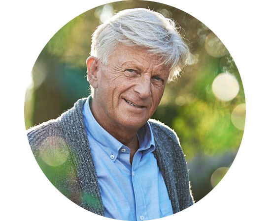 Elderly man outdoors wearing a blue shirt and gray cardigan with sunlight and greenery in the background.