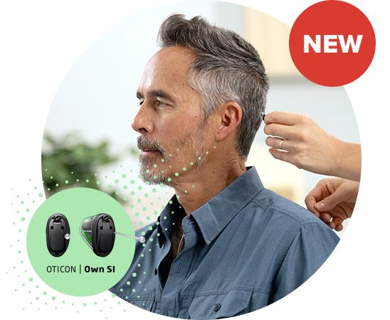 A person with short, salt-and-pepper hair sits in a hearing clinic, as an audiologist adjusts a discreet behind-the-ear Oticon Own SI hearing aid. A ’NEW’ badge and hearing aid images are displayed.